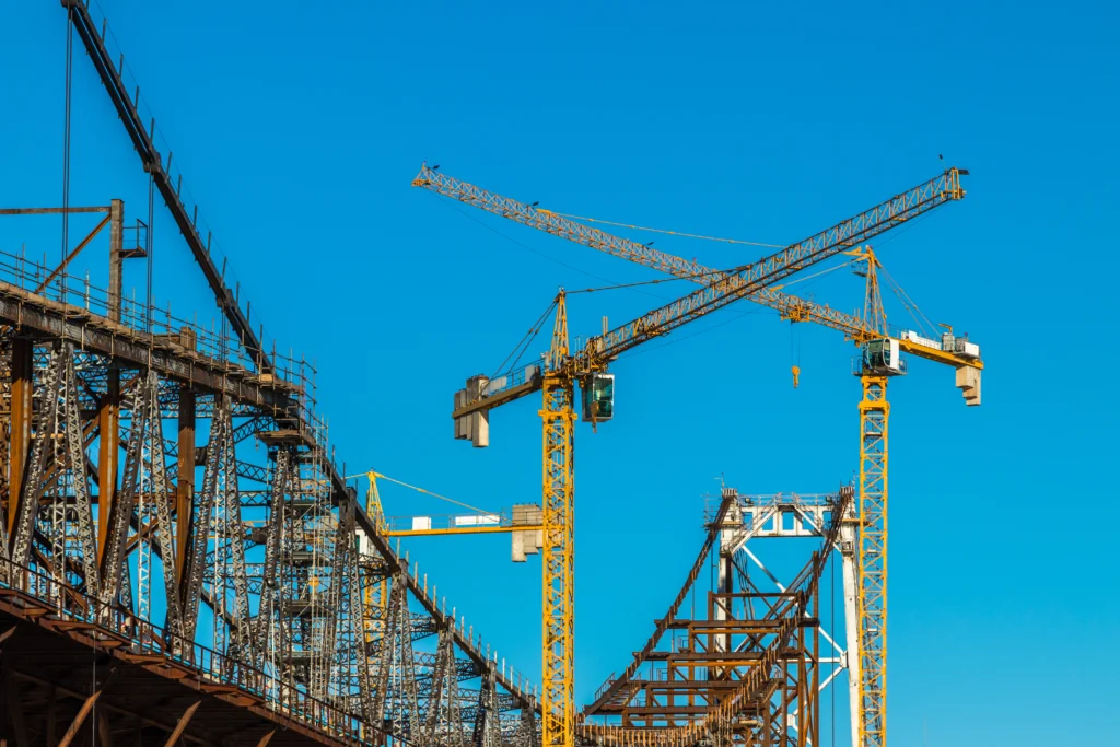 Chantier de construction du pont Hercílio Luz avec grues à tour, structures métalliques et travaux de génie civil sous un ciel bleu.
