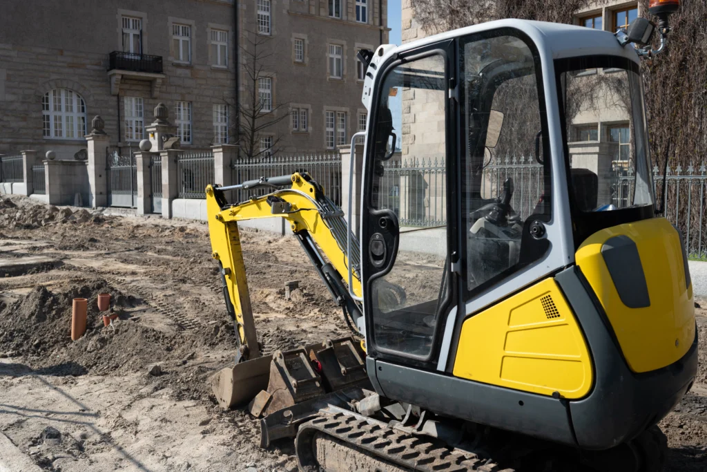 Mini-pelle hydraulique jaune sur un chantier urbain, utilisée pour les travaux de terrassement et la préparation de routes.